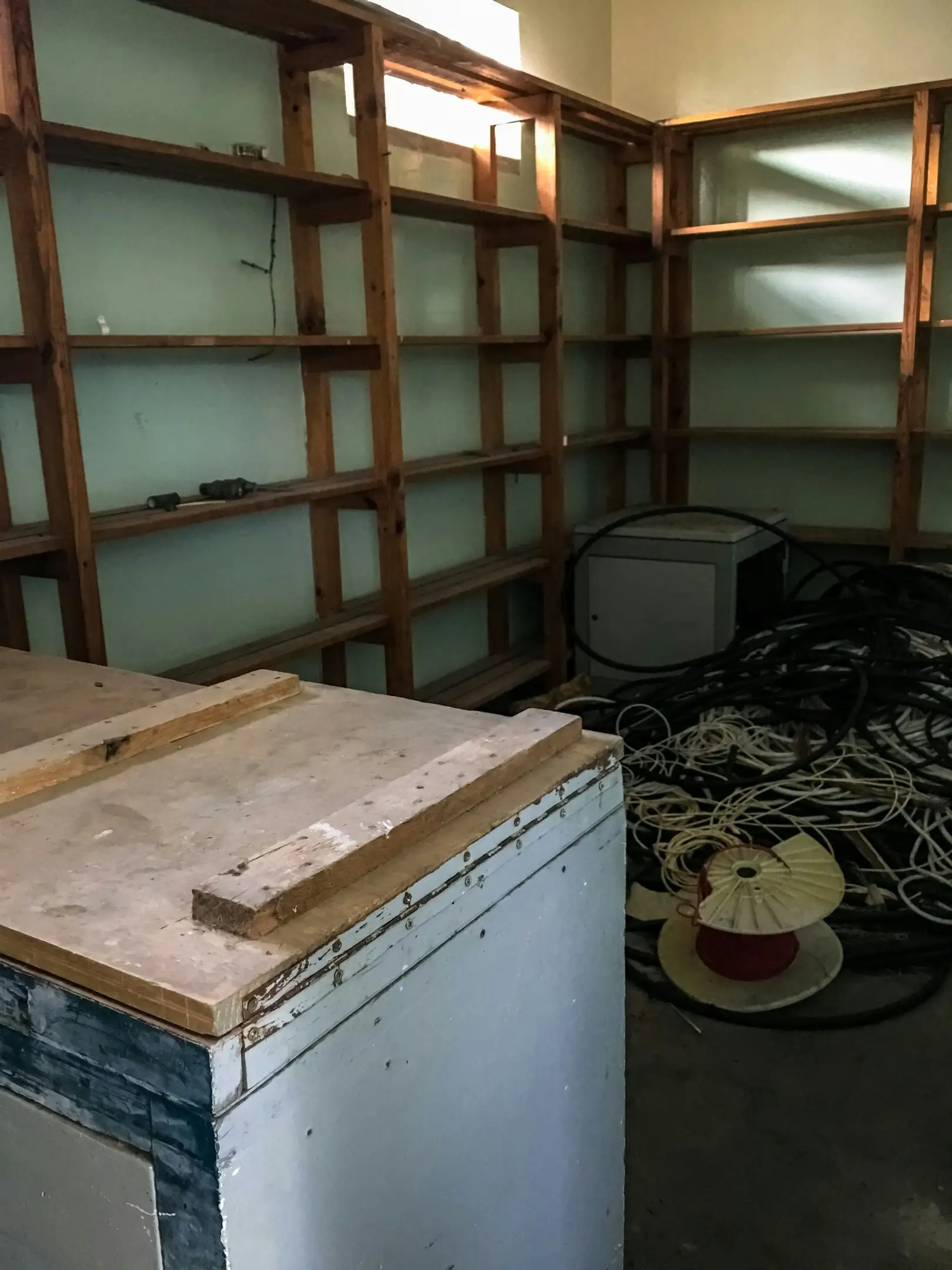 اتاقی نامرتب با کف پوشیده از کابل‌های برق قدیمی؛ دیوارهای سبز روشن قفسه‌های چوبی خالی را نشان می‌دهند.