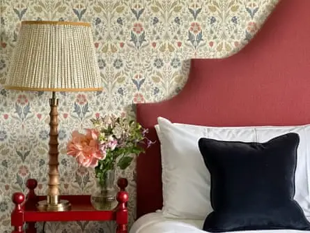 Close up of red hotel bed headboard, flowery wallpaper, dark cushion and red bedside table with lamp and vase of flowers