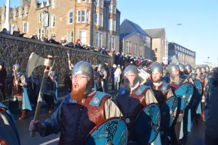 Men marching in daytime in Viking costumes and helmets, carrying axes and shields