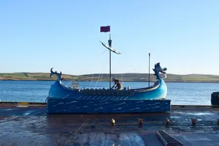 A man on a blue longship against a backdrop of sea and low hills